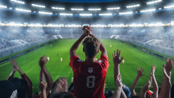 Backview Of Fans Cheer for Their Team on a Stadium During Soccer Championship Match. Teams Play, Crowds of Fans Celebrate Victory and Goal. Football Cup Tournament With Supporters Clapping On Tribune