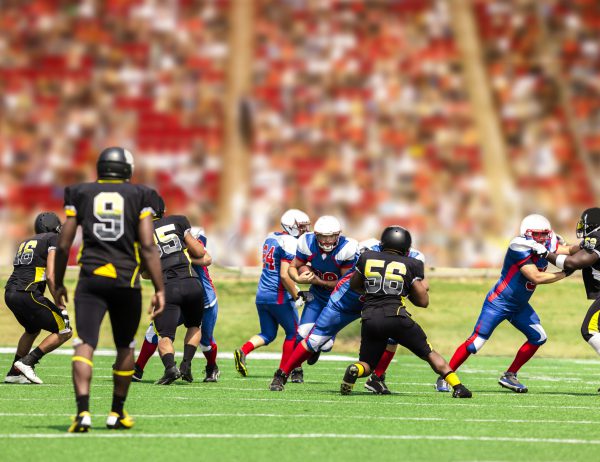 Semi-professional football team's running back carries the football to make a play. Defenders try to tackle him. Football field with a stadium full of unrecognizable fans in background.